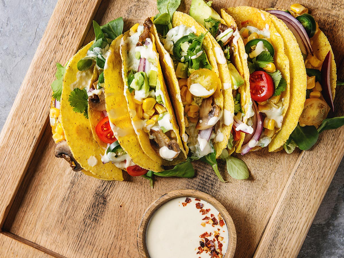 A spread of street tacos on a board with corn, mushrooms, tomatoes, and crema.